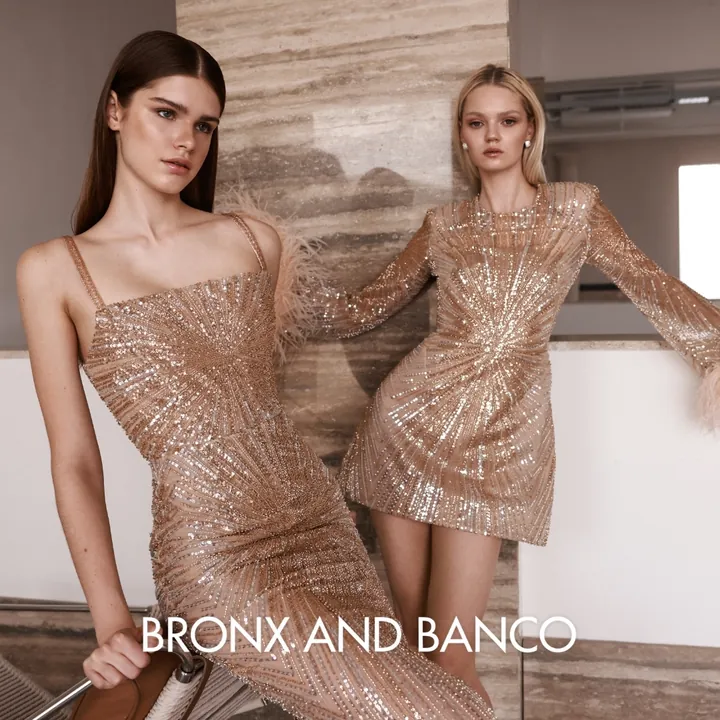Bronx and Banco ad: 2 models in beige sequin dresses with feather cuffs against a marble wall
