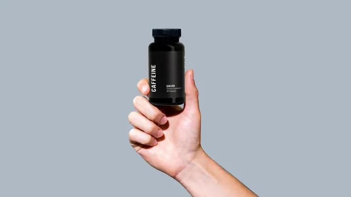 A hand holds up a bottle of Elm & Rye Caffeine capsules against a blue-gray backdrop