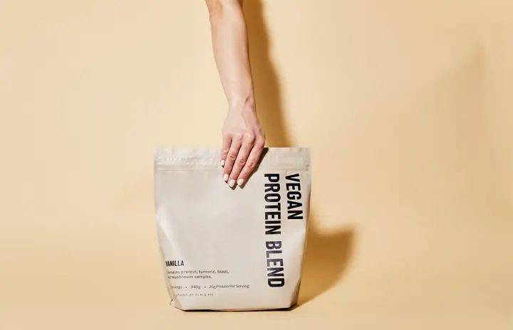 A hand lifts a pouch of Elm & Rye Vegan Protein Blend by its top edge against a peach backdrop