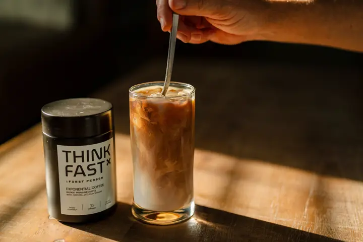 Iced coffee being stirred beside a jar of First Person Think Fast in low afternoon sun