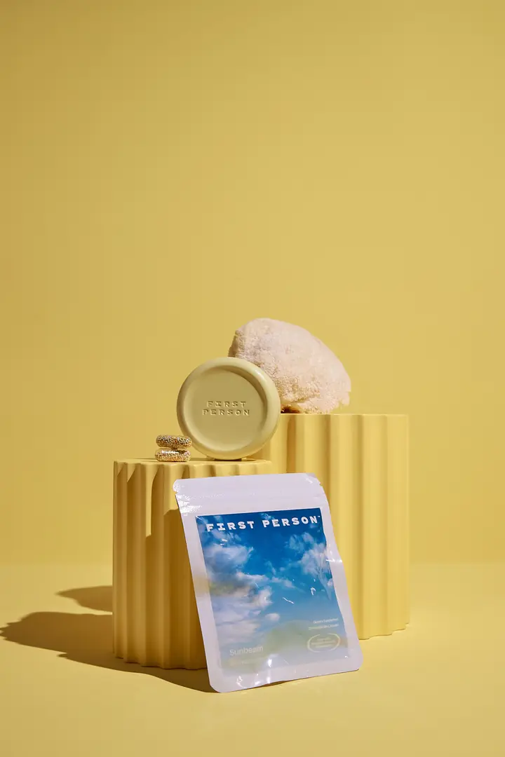 A First Person tin and supplement pouch on a yellow fluted pedestal beside a lion's mane mushroom