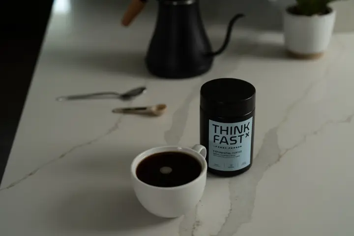 A jar of First Person Think Fast beside a cup of black coffee and a gooseneck kettle on marble