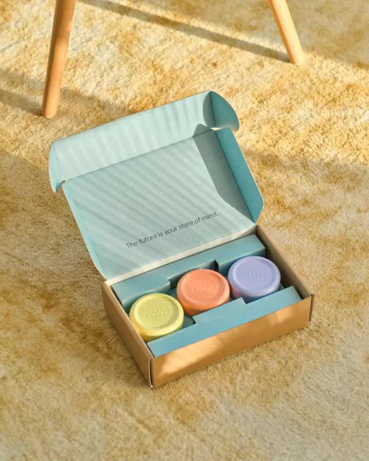 An open First Person mailer box holding 3 pastel supplement tins on a sunlit rug