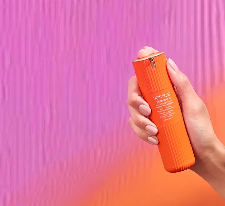 A hand holds an orange bottle of Lion Pose Unspotted serum against a pink-to-orange backdrop