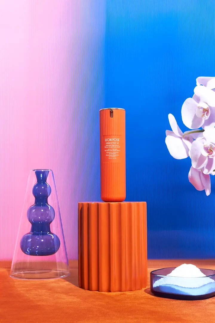 An orange bottle of Lion Pose Unspotted serum on a fluted pedestal beside a glass vase and an orchid