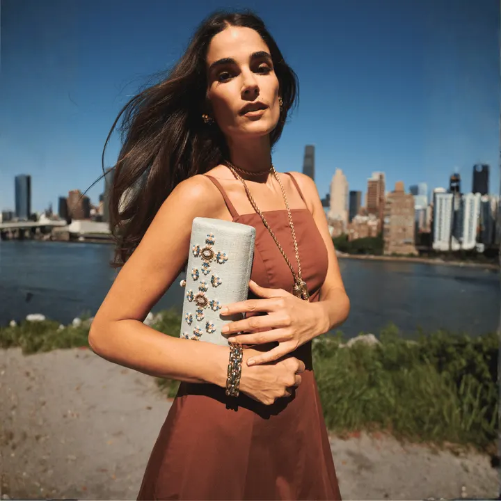A model in a rust Mestiza dress holds an embellished clutch, with the New York skyline behind her