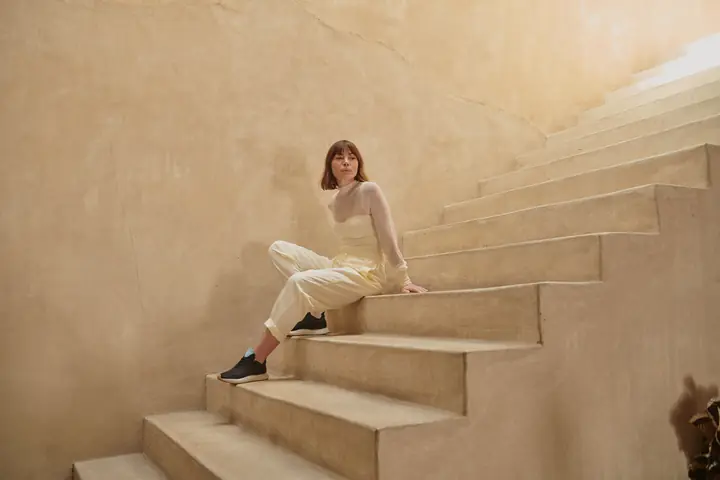 A model in cream knitwear and Saysh sneakers sitting on sunlit stone stairs