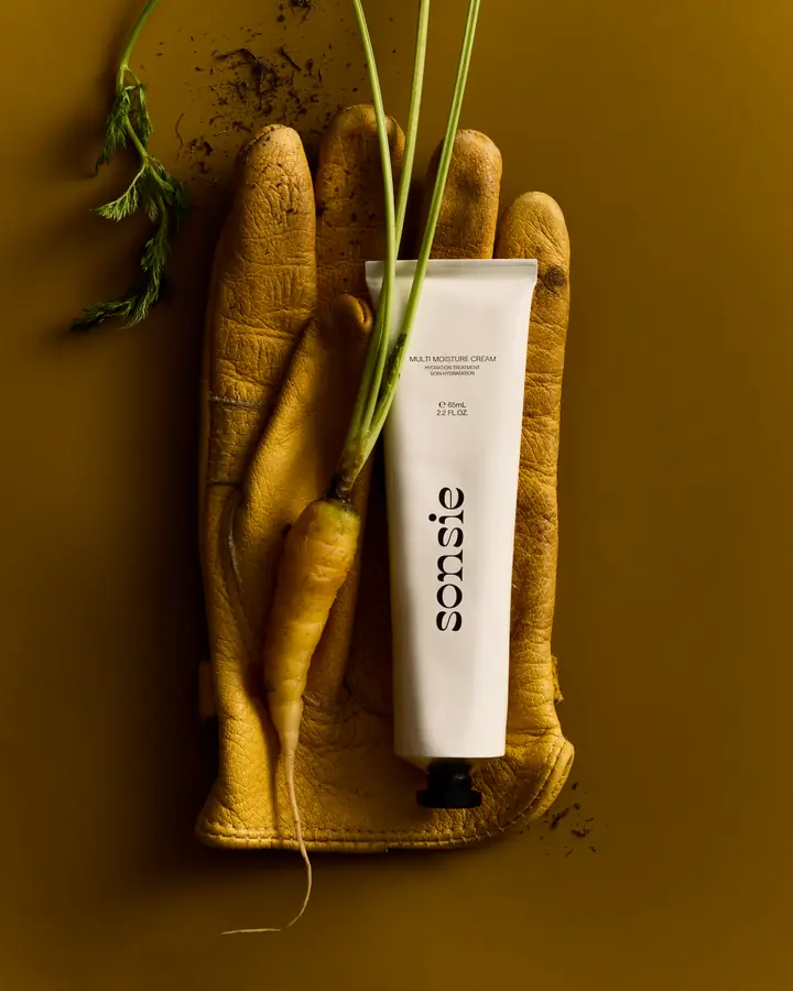 A tube of Sonsie Multi Moisture Cream resting on a yellow gardening glove with fresh carrots