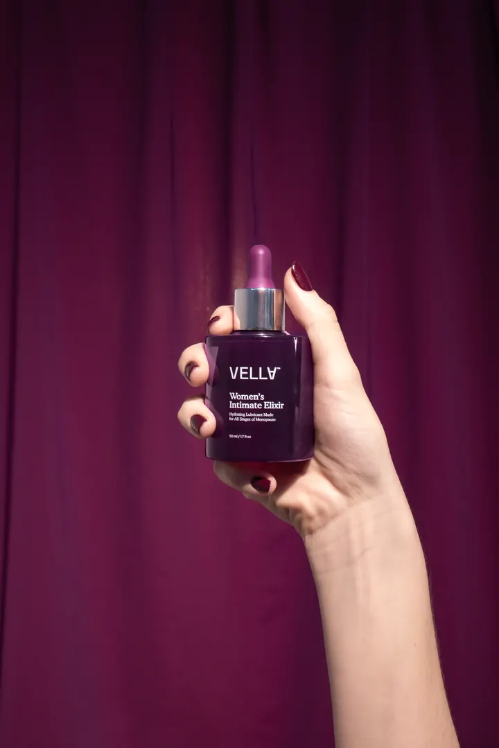 A hand holds up a bottle of Vella Women’s Intimate Elixir against a plum velvet curtain