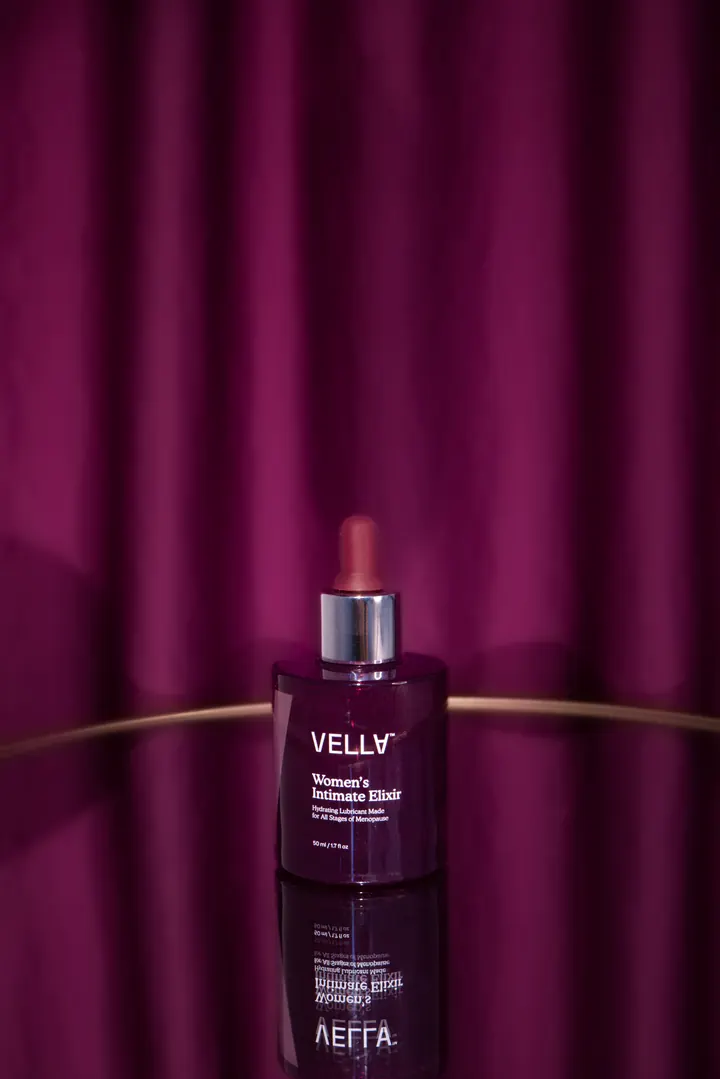A bottle of Vella Women’s Intimate Elixir on a gold-rimmed stage, reflected in the floor, before a plum curtain