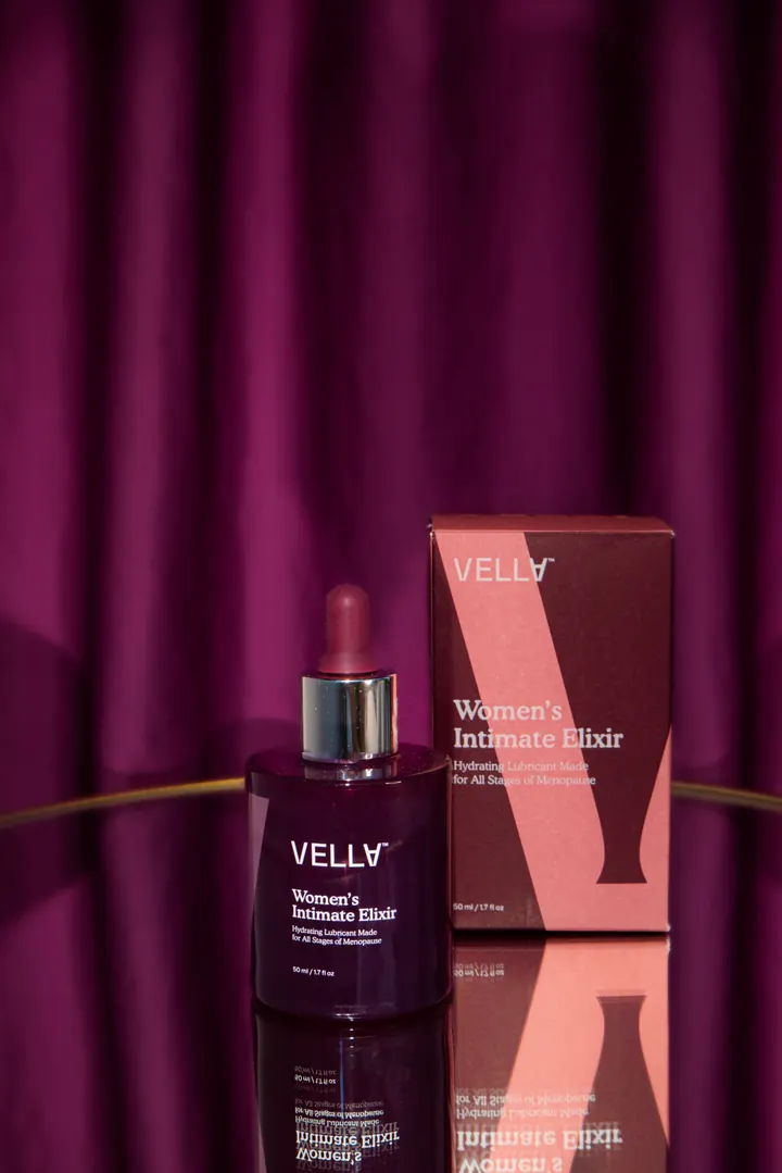 A bottle of Vella Women’s Intimate Elixir beside its pink box on a small stage in front of a plum curtain